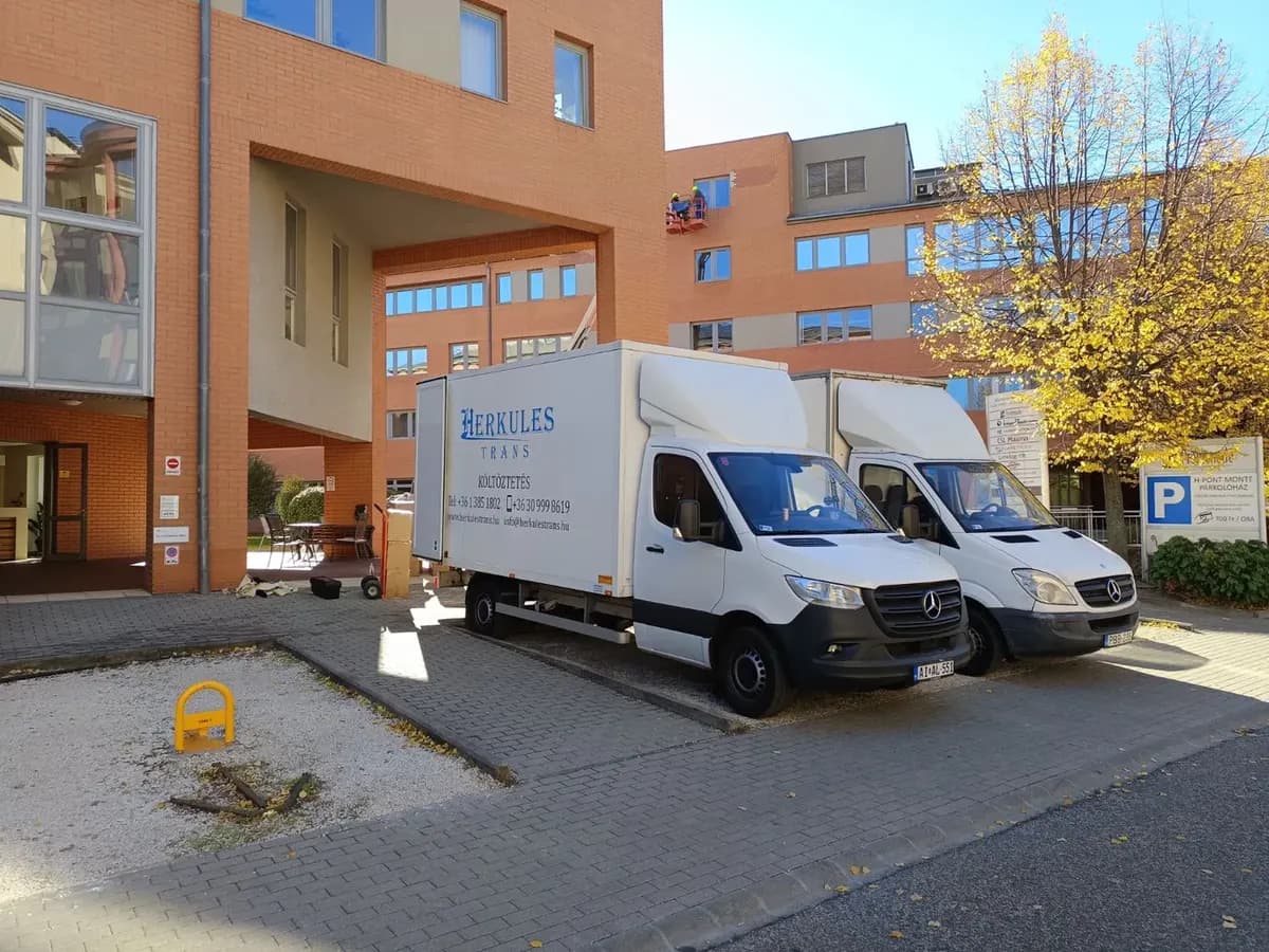 Két fehér Herkules-Trans költöztető teherautó egy budapesti irodaépület udvarán, őszi fák között, mellettük dobozok és kézikocsi